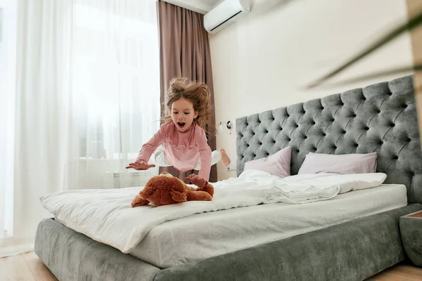 A little barefoot girl enjoying jumping down on her teddybear with her hair back on a bed — Stock Photo, Image
