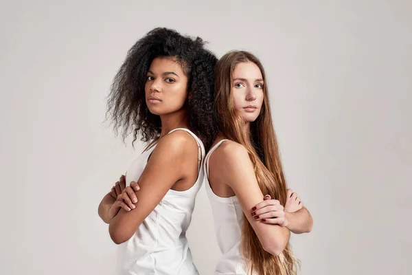 Ritratto di due giovani donne diverse che indossano camicie bianche guardando la macchina fotografica mentre stanno schiena contro schiena isolate su sfondo grigio — Foto Stock
