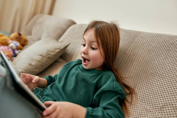 Uma menina pequena bonito ter um bom tempo jogando videogames em het tablet gritando emocionalmente sentado em um sofá — Fotografia de Stock