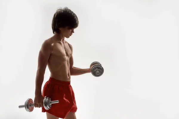 Little sportive boy child with muscular body exercising, showing his muscles, lifting weights while standing isolated over white background — Stockfoto