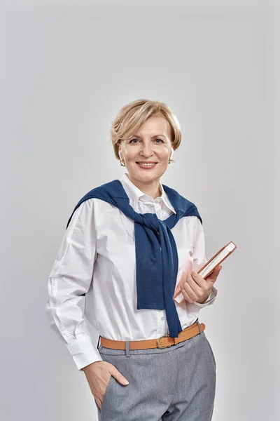 Portrait of elegant middle aged caucasian woman wearing business attire smiling at camera, holding her notebook while standing isolated over grey background —  Fotos de Stock