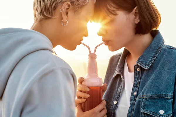 Szczęśliwa młoda lesbijska para mając romantyczny moment, pijąc z jednej szklanej butelki ze słomką, dwie kobiety cieszące się zimnym napojem w letni dzień na świeżym powietrzu — Zdjęcie stockowe