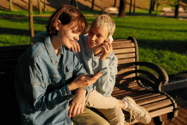 Δύο χαρούμενες γυναίκες που χρησιμοποιούν smartphone, κάθονται στον πάγκο στο πάρκο της πόλης. Λεσβιακό ζευγάρι που κρατάει τα τηλέφωνά του ενώ περνάει χρόνο μαζί στην ύπαιθρο — Φωτογραφία Αρχείου