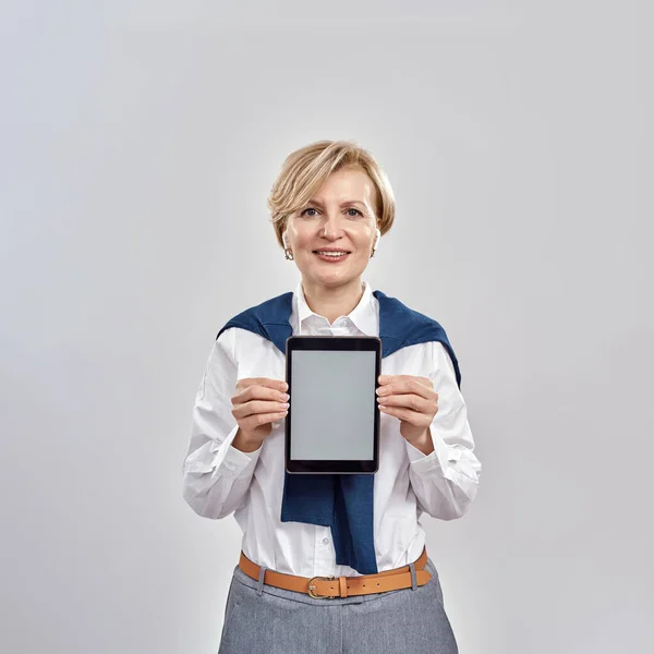 Ritratto di elegante donna caucasica di mezza età che indossa abiti da lavoro sorridente alla macchina fotografica, tenendo tablet pc con schermo vuoto, applicazione pubblicitaria mentre in piedi isolato su sfondo grigio — Foto Stock