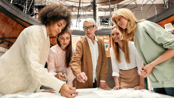 Un grupo de mujeres de diferentes edades y nacionalidades hablando juntas y paradas alrededor de una mesa durante una lluvia de ideas — Foto de Stock