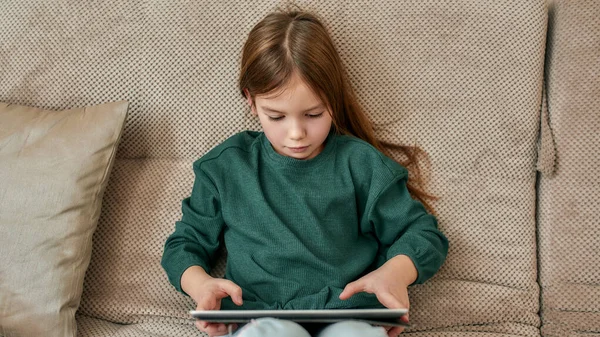 Uma menina batendo em uma tela de seu tablet sentado em um sofá — Fotografia de Stock
