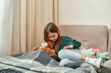 Ekoseli kanepeye uzanmış tatlı küçük bir kız ve parlak bir odada oyuncak ayısı.