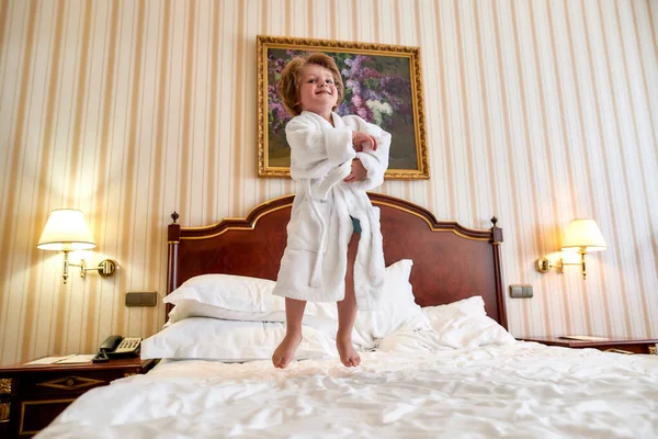 Відкрийте для себе щастя. Веселий хлопчик у білому халаті розважається, стрибає на білому ліжку в готельному номері. Дитина сміється і стрибає — стокове фото