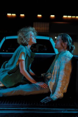 Farklı cinsiyetlerden bir grup yakışıklı beyaz arkadaş bir otoparkın dışında açık bir arabanın bagajında birbirlerine bakıyorlar.