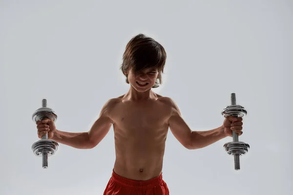 Little sportive boy child with muscular body looking emotional while exercising, lifting weights, standing isolated over grey background — Stock Photo, Image