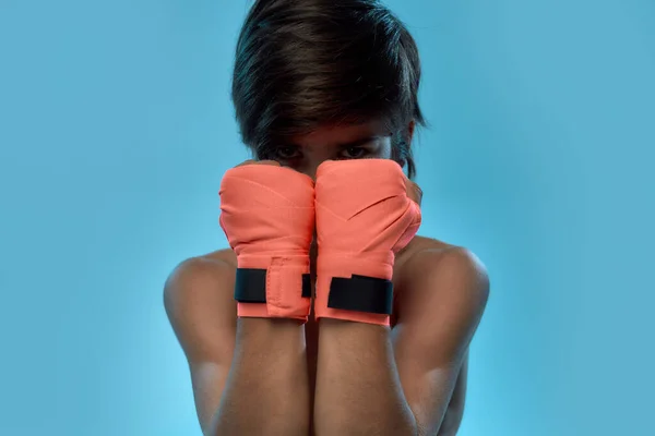 Close up retrato de menino boxeador com bandagem de boxe nas mãos, olhando para a câmera, cobrindo o rosto com as mãos enquanto posando, de pé isolado sobre fundo azul — Fotografia de Stock