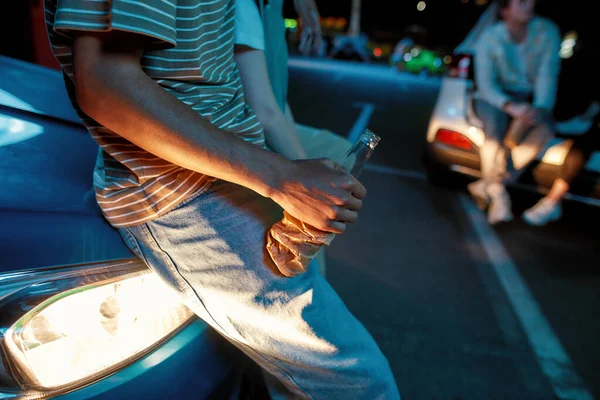 A close up of a bottle of beer wrapped in paper bag holding by a young man leaning against a car outside on a parking site with friends around and a led screen on a background — Stock Photo, Image