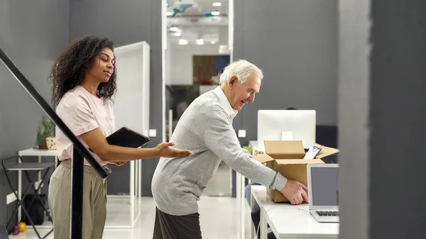 Friendly female company executive supporting, welcoming new employee aged man, senior intern putting cardboard box with belongings on the table — Stock Photo, Image