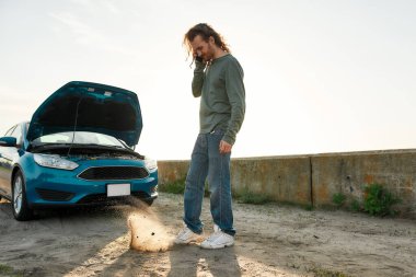 Kaputu açık duran bozuk arabasının yanında duran genç adamın araba servisi, yardım ya da çekici ile telefonda konuşurken tam boy görüntüsü.