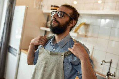 Genç adam, gözlüklü profesyonel aşçı yemek hazırlarken önlüğünü giyiyor, mutfakta duruyor. Ev konseptinde aşçılık