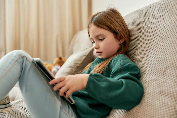 Uma garotinha fofa ocupada com seu tablet olhando na tela enquanto sentada em um sofá — Fotografia de Stock