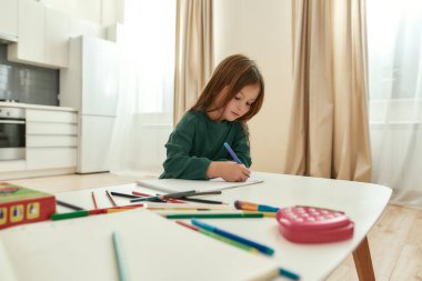 Küçük tatlı bir kız kalemleriyle deftere not alıyor ve spacy mutfağındaki bir masada oturuyor.