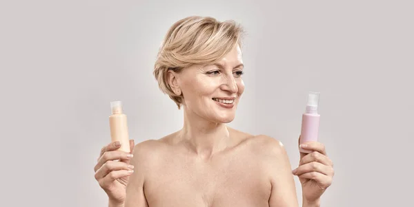 Retrato de bela mulher de meia-idade segurando duas garrafas de produtos cosméticos diferentes e escolhendo o que aplicar ao posar isolado sobre fundo cinza — Fotografia de Stock