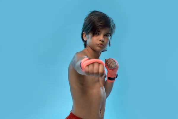 Portret małego chłopca boksera z bandażem bokserskim na rękach, patrzącego w kamerę, uderzającego podczas pozowania, stojącego na niebieskim tle — Zdjęcie stockowe
