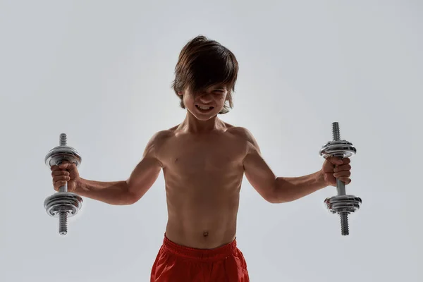 Little sportive boy child with muscular body looking emotional while exercising, lifting weights, standing isolated over grey background — Stock Photo, Image