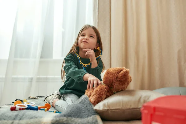 Malá holka hrající si na doktora se stetoskopem na hračky, která má plyšového medvídka jako pacienta, který se rozhoduje, co dělat, zatímco sedí na pohovce ve spacy Room — Stock fotografie