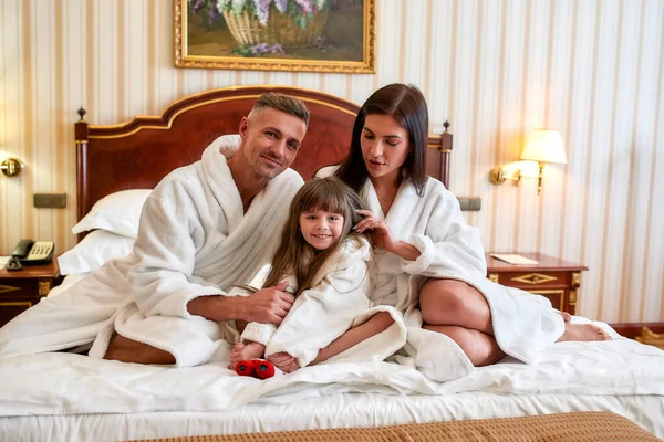 Це чудове місце, щоб бути. Кавказькі батьки і дочка в білому халаті сидять на ліжку в розкішному готельному номері. Люди, подорожі, туризм, відпустка. — стокове фото