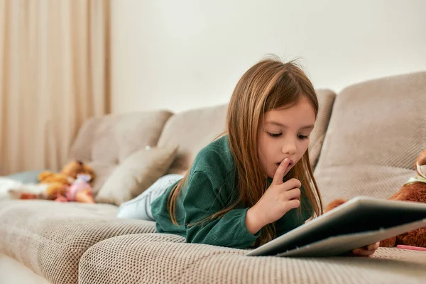 A little cute girl lying on her stomach on a sofa holding a tablet putting a point finger to her opened mouth — Stock Photo, Image