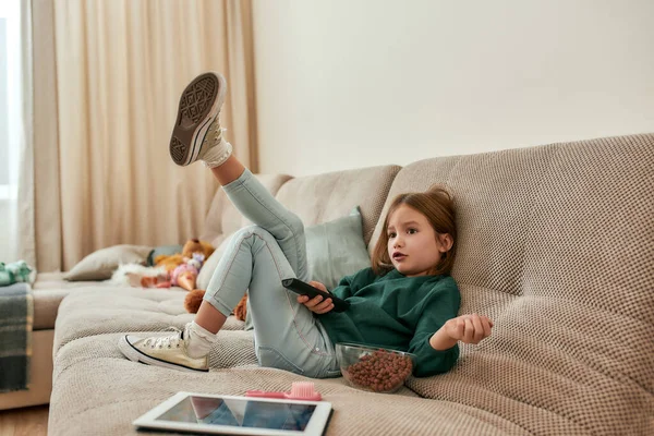 Una linda niña pequeña preguntándose qué ver sosteniendo un televisor remoto sentado en un sofá en una habitación grande —  Fotos de Stock