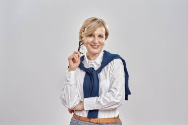 Kafkasyalı zarif bir kadının portresi iş elbisesi giyiyor, gözlüklerini tutuyor, kameraya gülümsüyor ve gri arka planda tek başına dikiliyor.