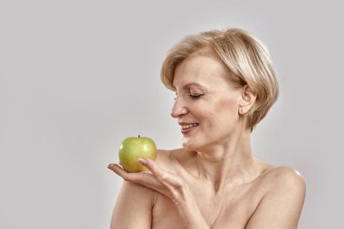 Güzel orta yaşlı kadın yeşil elmaya bakıyor, gri arka planda soyutlanmış poz veriyor.
