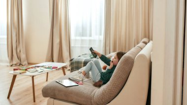 Küçük bir kız elinde uzaktan kumandayla kanepede oturmuş televizyon seyrediyor tabletini ve oyuncaklarını etrafta gezdiriyor.