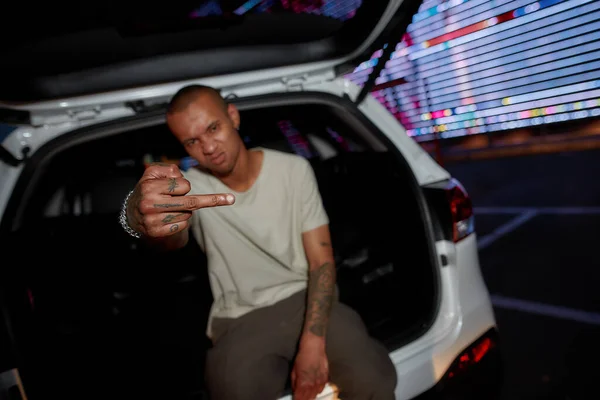 A dark-skinned tattooed well-dressed guy looking into a camera with a middle finger out sitting in an opened car trunk on a parking site with a led screen behind