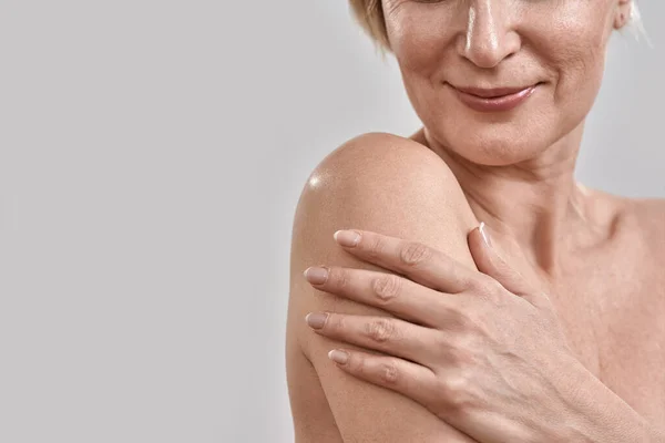 Крупним планом знімок руки і плеча красивої жінки середнього віку, яка торкається її шкіри, позує ізольовано на сірому фоні. Краса, концепція скінарію — стокове фото