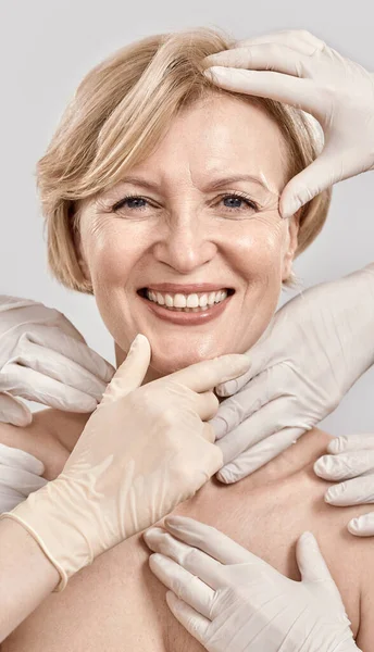 Close up retrato de mulher de meia-idade atraente sorrindo para a câmera. Mãos de esteticista em luvas verificando a pele feminina do rosto isolado contra fundo cinza — Fotografia de Stock