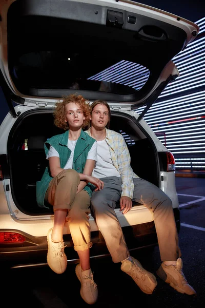 Skupina dvou mladých dobře oblečených bílých přátel různých pohlaví hledících do kamery sedící v otevřeném kufru auta venku na parkovišti s LED obrazovkou za — Stock fotografie