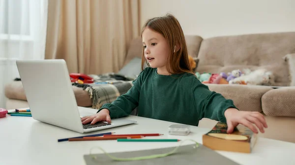 Uma menina bonito arrumando um livro para voltar ao seu laptop sentado sozinho em uma mesa em um grande quarto brilhante sozinho durante a educação à distância — Fotografia de Stock