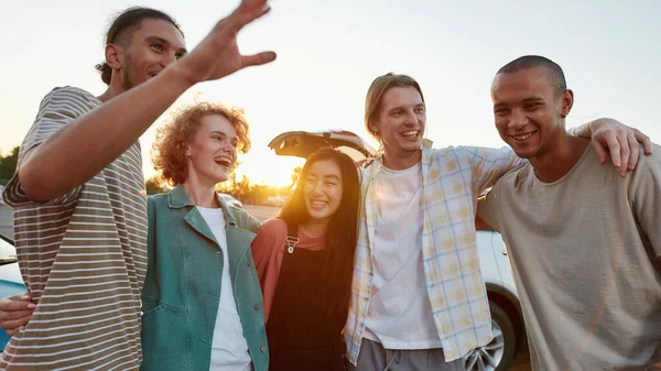 Pięciu przypadkowo ubranych przyjaciół różnych narodowości przytulających się do siebie, dobrze się razem bawiących na zewnątrz, śmiejących się i uśmiechających swoim samochodem na tle. — Zdjęcie stockowe