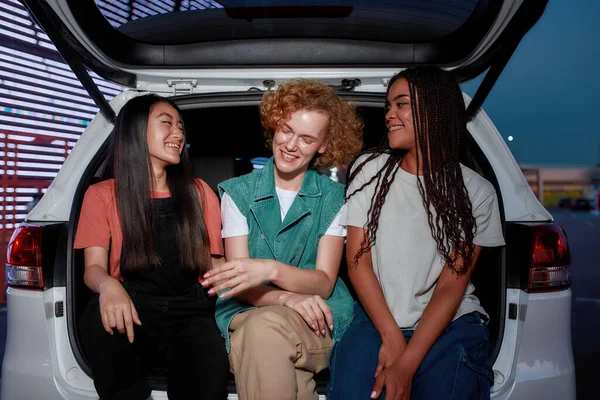 Un primer plano de un grupo de tres jóvenes mujeres bien vestidas de diferentes nacionalidades sentadas en un maletero abierto afuera en un sitio de estacionamiento riendo juntas — Foto de Stock