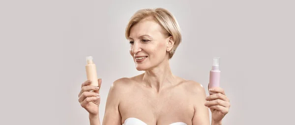 Portrait of beautiful middle aged woman holding two bottles of different cosmetic skincare products and choosing what to apply while posing isolated over grey background — Stock Photo, Image