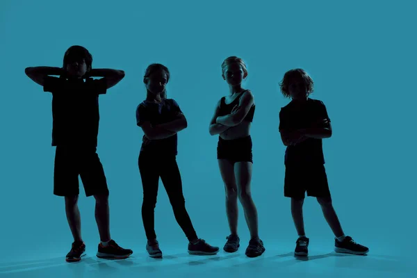 Tiro de comprimento total silhueta de quatro crianças esportivas olhando para a câmera enquanto posam, em pé isolado sobre o fundo azul — Fotografia de Stock
