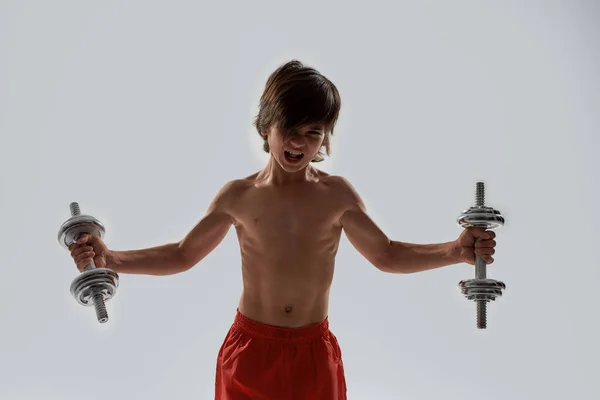 Little sportive boy child with muscular body looking emotional while exercising, lifting weights, standing isolated over grey background — Stock Photo, Image