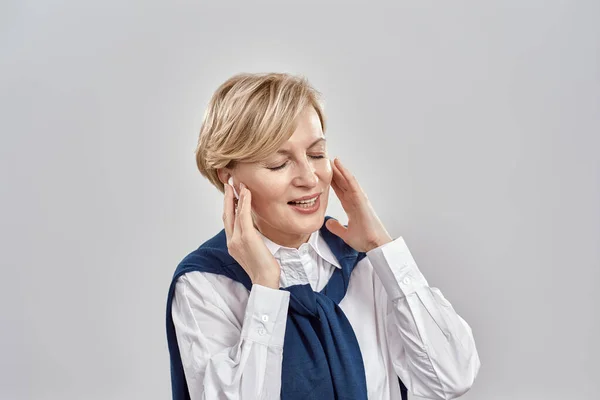 Retrato de mulher branca de meia-idade elegante vestindo trajes de negócios, ajustando seus fones de ouvido, apreciando o som com os olhos fechados enquanto posando isolado sobre fundo cinza — Fotografia de Stock