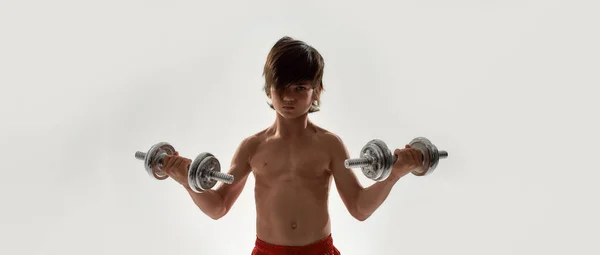 Little sportive boy child with muscular body looking focused at camera, lifting weights while standing isolated over white background — Stock Photo, Image