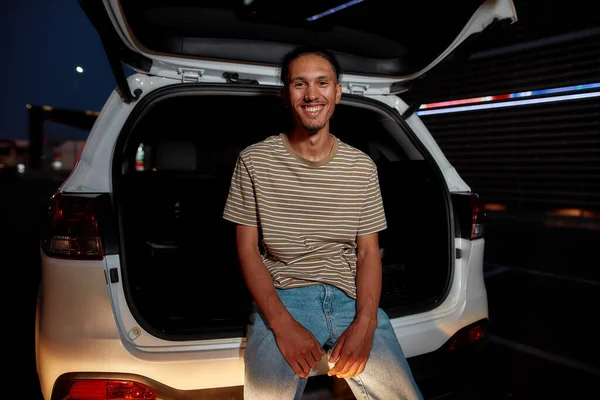 A commonly dressed dark-skinned young latina man with a piercing smiling looking into a camera sitting in an opened car trunk outside on a parking site with a led screen behind him