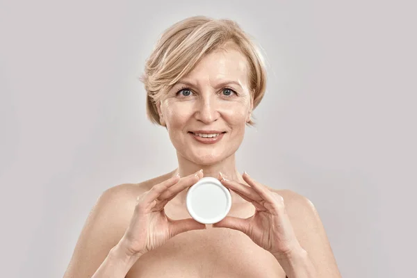 Portrait of beautiful middle aged woman smiling at camera, holding moisturizing facial cream while posing isolated over grey background — Stock Photo, Image
