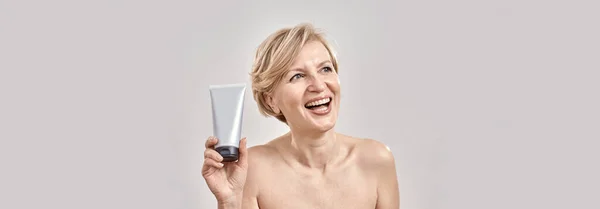 Beautiful middle aged woman looking happy, holding, advertising cosmetic skincare bodycare product while posing isolated over grey background — Stock Photo, Image