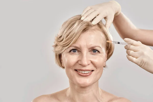 Retrato de bela mulher de meia-idade nua sorrindo para a câmera enquanto ela recebe injeção em seu rosto, posando isolado contra fundo cinza — Fotografia de Stock