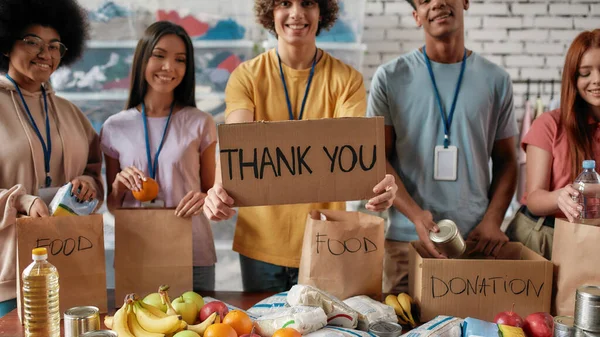 Recortado tiro de grupo de jóvenes voluntarios diversos embalaje de alimentos y bebidas donación, Guy celebración de la tarjeta con letras Gracias, Frutas, latas y paquetes sobre la mesa — Foto de Stock