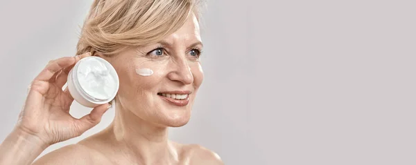Portrait of beautiful middle aged woman smiling aside, holding cream jar while posing with cream applied on her face isolated over grey background — Stock Photo, Image