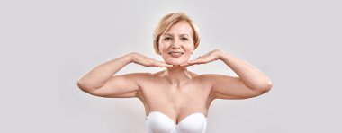 Cilt bakımından memnun orta yaşlı güzel bir kadının portresi. Yüzünün yanında el ele tutuşuyor, gri arka planda izole edilmiş kameraya gülümsüyor. Güzellik konsepti